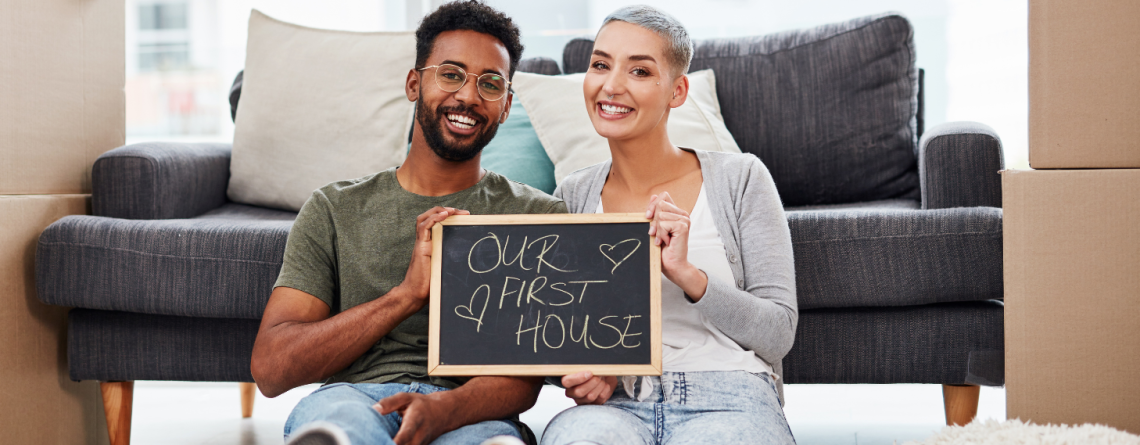 Couple smiling and holding a chalk board with "Our First Home" written on it.