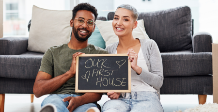 Couple smiling and holding a chalk board with "Our First Home" written on it.