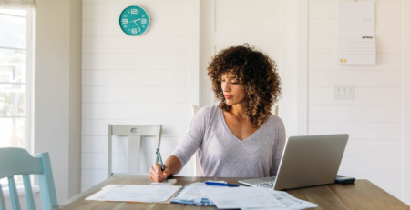 A woman focused on financial spring cleaning, reviewing documents and using a laptop at a sunlit table with an April calendar in the background.
