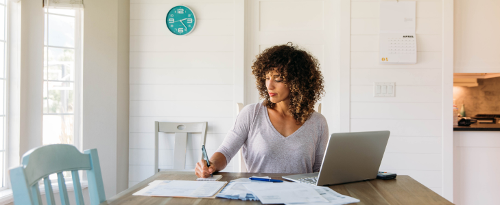 A woman focused on financial spring cleaning, reviewing documents and using a laptop at a sunlit table with an April calendar in the background.