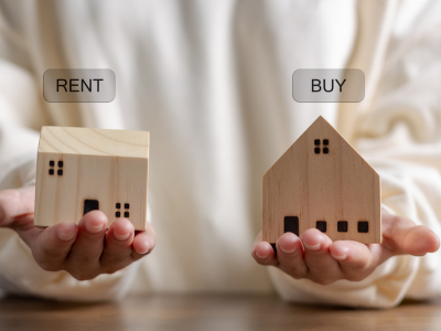 A person holding wooden house models labeled 'Rent' and 'Buy' to compare housing options in Missouri and Illinois.