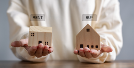 A person holding wooden house models labeled 'Rent' and 'Buy' to compare housing options in Missouri and Illinois.