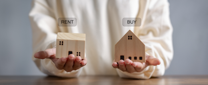 A person holding wooden house models labeled 'Rent' and 'Buy' to compare housing options in Missouri and Illinois.