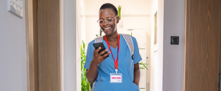 A smiling healthcare worker in blue scrubs looking at her phone as she walks through the front door of her home, illustrating mortgage loan options for community heroes at Scott Credit Union.