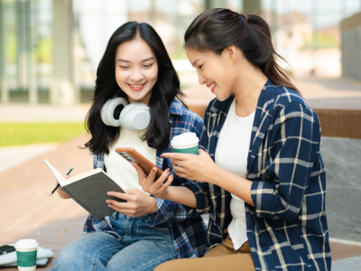 Two college students sitting outdoors on campus, holding coffee and reviewing mobile banking on a smartphone with an open notebook.