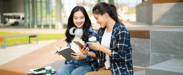 Two college students sitting outdoors on campus, holding coffee and reviewing mobile banking on a smartphone with an open notebook.