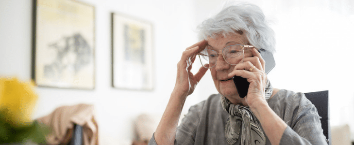 Concerned older woman holding her head while talking on a smartphone during an AI voice scam call.