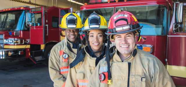 Three smiling firefighters in uniform outside a fire station bay with fire trucks, representing Scott Credit Union's Community Heroes Loan Program.