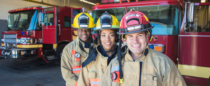 Three smiling firefighters in uniform outside a fire station bay with fire trucks, representing Scott Credit Union's Community Heroes Loan Program.